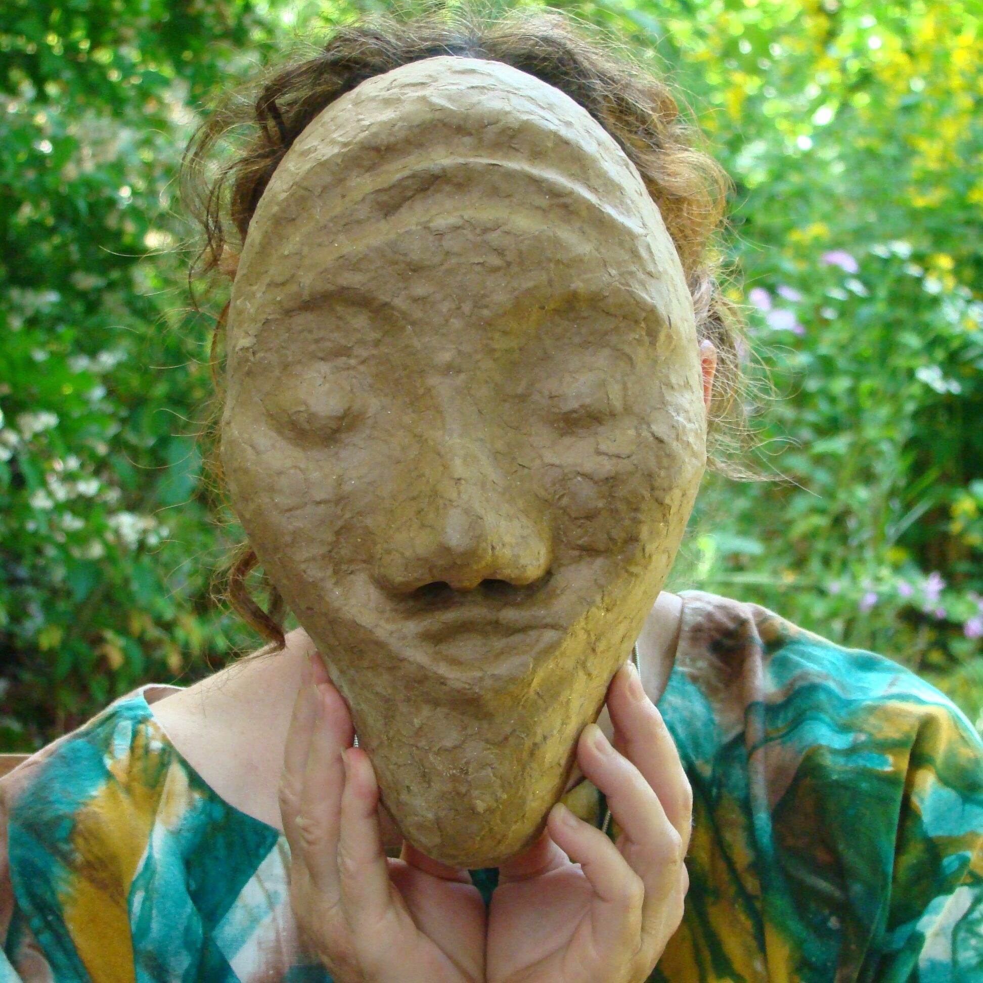 Person hält unbemalte Maske vor ihr Gesicht, Ausdruck der Maske is tverspielt.